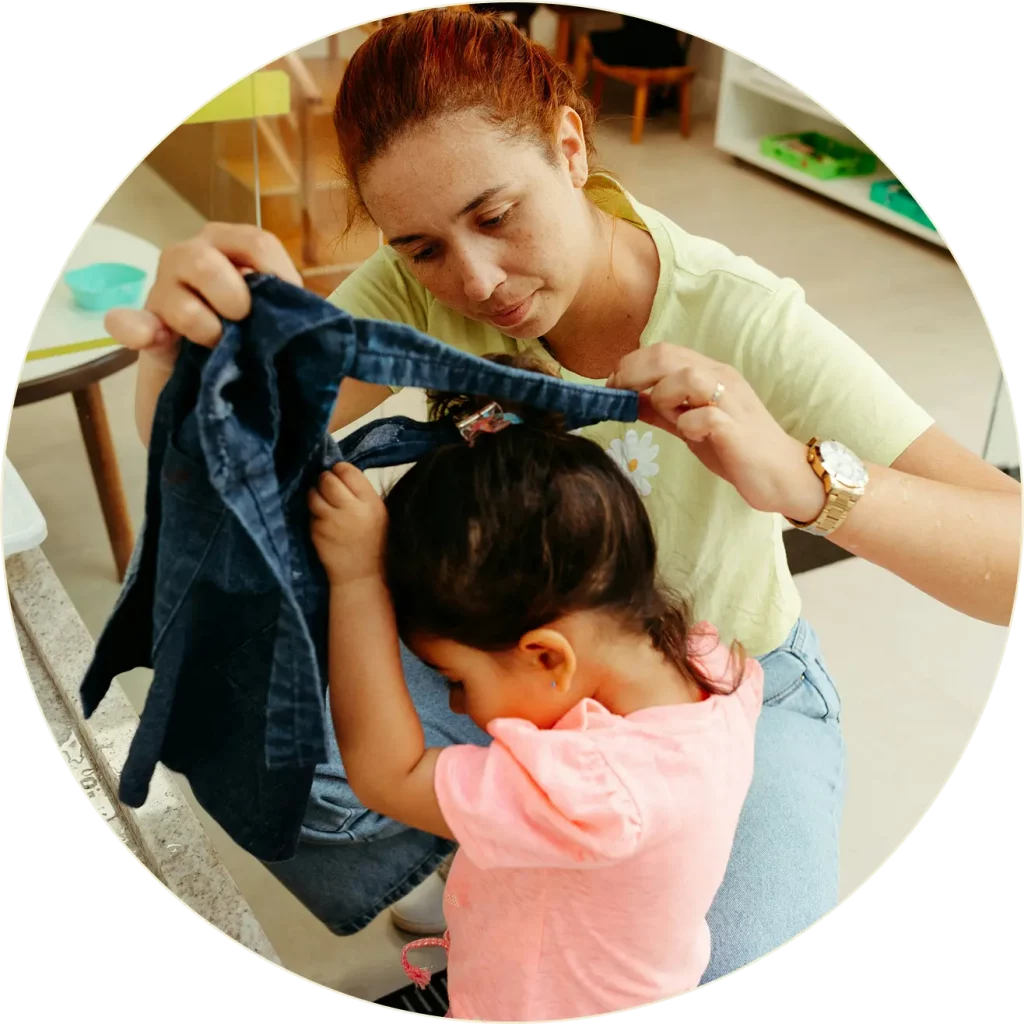 Une femme aide une jeune fille à enfiler une veste en jean. La femme est assise et tient la veste tandis que la jeune fille, qui porte une chemise rose, penche la tête pour passer ses bras dans les manches. Elles sont à l'intérieur.