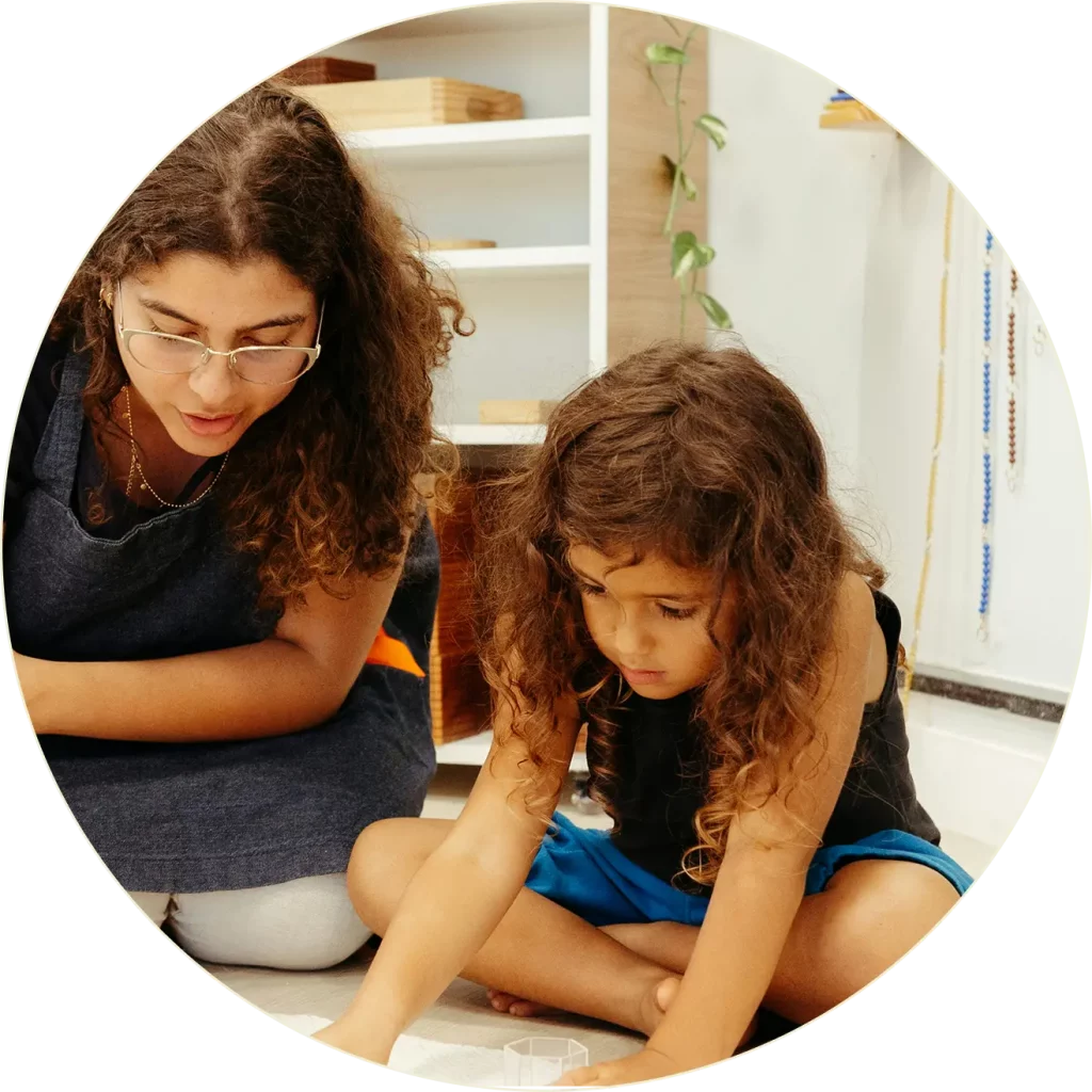 Une femme adulte et un jeune enfant aux cheveux bouclés sont assis par terre, étroitement engagés dans une activité éducative ou créative à l'intérieur. Des étagères contenant du matériel sont visibles à l'arrière-plan.