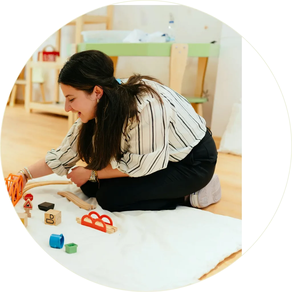 Une femme aux longs cheveux noirs est agenouillée sur le sol, souriante, alors qu'elle arrange des jouets en bois colorés sur un tapis blanc dans une pièce lumineuse avec des planchers en bois.