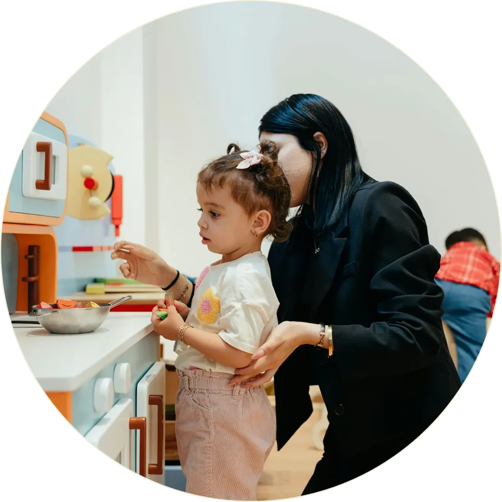 Une femme aide un jeune enfant à jouer avec un jouet de cuisine, en guidant ses mains pendant que l'enfant se tient devant une cuisinière et des ustensiles. Un autre enfant est visible à l'arrière-plan.