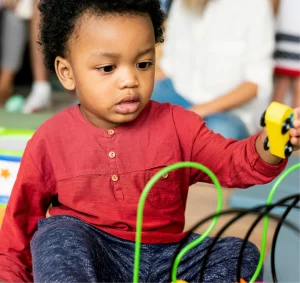 Toddler playing with a bead maze and a yellow toy car, focused expression.